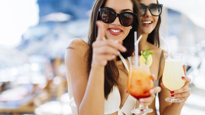 Two women in bikinis and sunglasses sipping cocktails by the pool