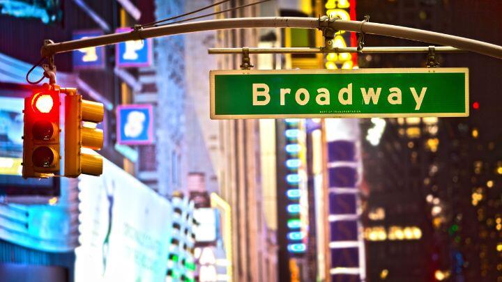 Green Broadway street sign next to a red traffic light, against abstract backdrop of lights and buildings