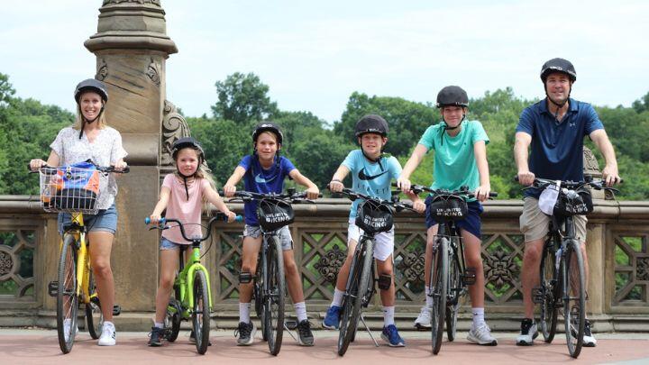 Family of mum, dad and four kids face the camera on bikes, in cycle helmets, against a bridge in Central Park