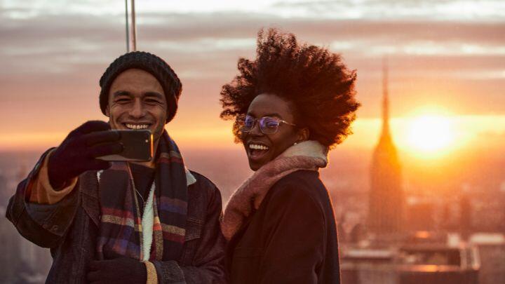 Man and woman with windswept hair take photo from high point against sunset view of Empire State Building