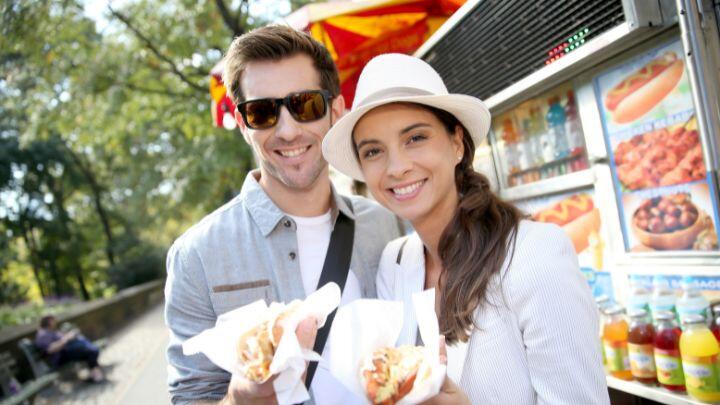 Man in sunglasses and lady in hat holding hot dogs in front of a hot dog stand