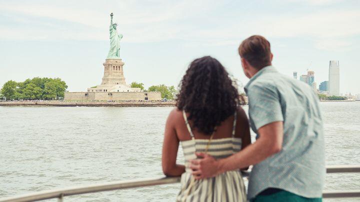 Rear view of couple, man with his arm around woman's waist, looking at a distant Statue of Liberty
