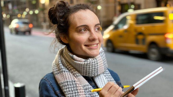 Happy woman with scarf holding drawing pad and pencil gazes off camera, with yellow cab background
