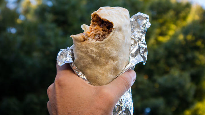 Hand holding a large, foil-wrapped burrito with a bite out of it