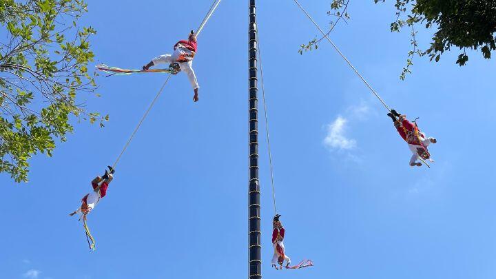 Voladores (flying men) spin upside down on ropes around a tall pole against a blue sky