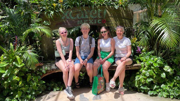 Smiling family of older kids on bench surrounded by tropical plants, under Xcaret sign