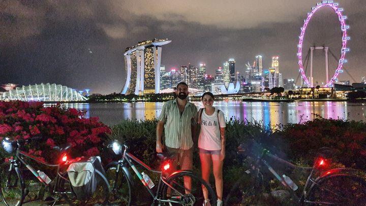 Couple pose for picture next to bicycles in front of Singapore night skyline reflecting colours into the water