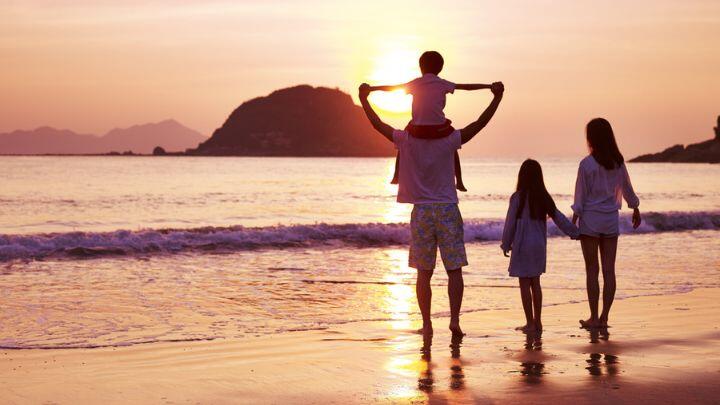 Family of four on beach, one kid on dad's shoulders, looking to sea, silhouetted against sunset