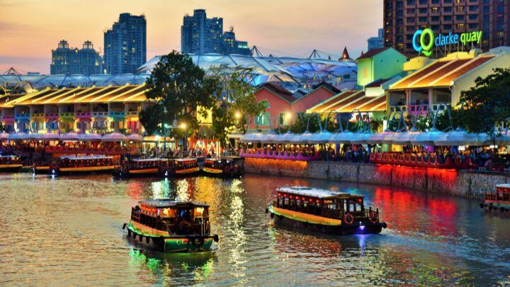 Brightly lit buildings reflect colour into a river containing two traditional boats - Singapore