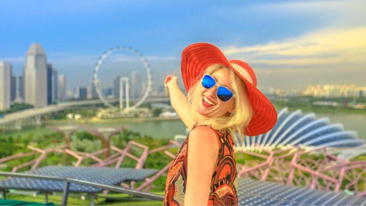 Smiling woman in sunglasses and red sunhat gestures towards sunny Singapore panorama