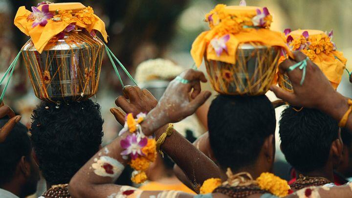 Back of the heads of 3 Hindu devotees carrying metal pots adorned with yellow cloths and flowers