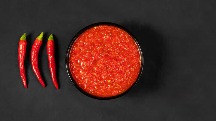 Bowl of bright-red samba chili sauce with three chili peppers next to it.