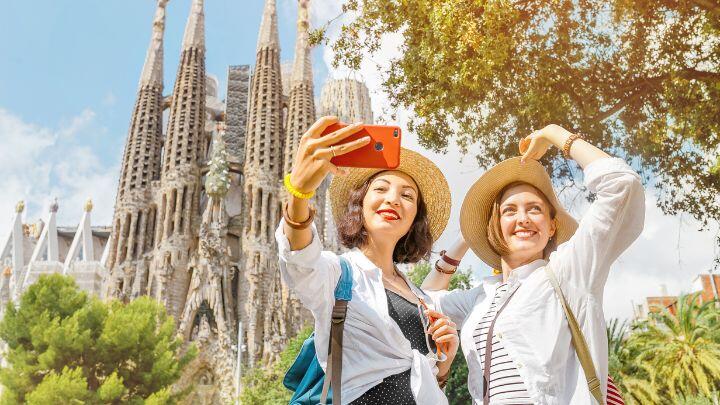 Two ladies in straw hats take a selfie in the sunshine in front of Sagrada Familia and trees