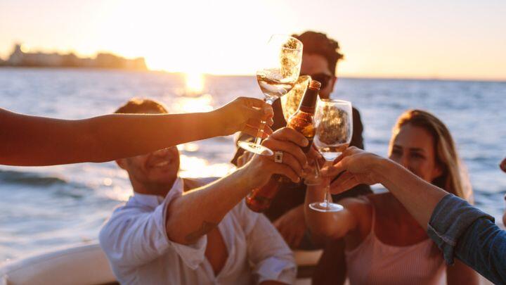 Group of adults raise a toast with wine glasses against backdrop of sea at sunset